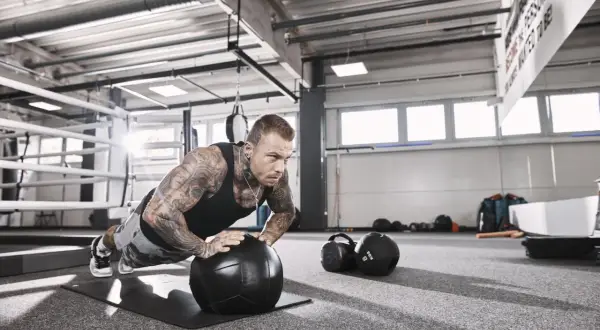 Mann mit Tattoos macht Liegestütze auf einem Medizinball, fokussiert auf sein hypertrophie training. Im Hintergrund ein Boxring und Trainingsgeräte.