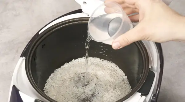 Beim Reis kochen wie viel Wasser? Eine Hand gießt Wasser in den Reiskocher über den Reis.