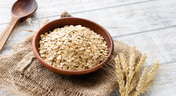 Schüssel mit Haferflocken, die für ihren guten Haferflocken proteingehalt bekannt sind, daneben eine Holzschale und Weizenähren.