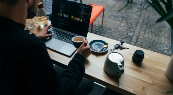 Mężczyzna pracuje na laptopie, popijając kawę. Widać kod na ekranie, klucze i aparat. To przykład multitasking co to znaczy być produktywnym.