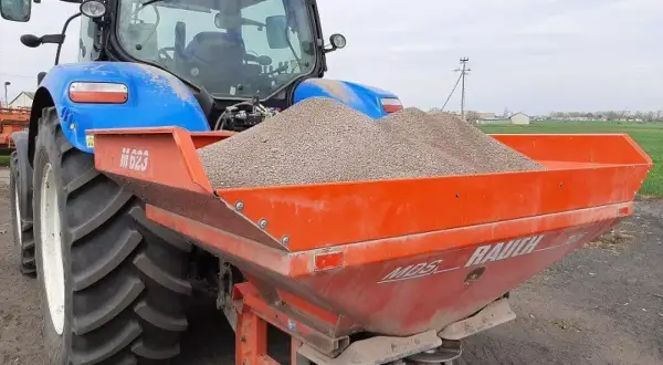 Niebieski traktor z przyczepionym rozsiewaczem napełnionym wapnem, gotowy do pracy na polu.
