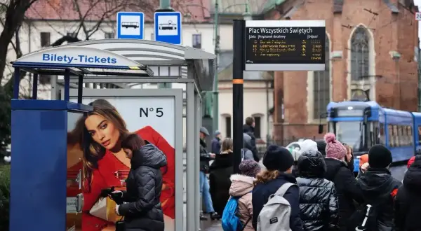 Ludzie czekają na przystanku Plac Wszystkich Świętych. Obok automatu z biletami miesięcznymi wisi tablica z rozkładem jazdy.
