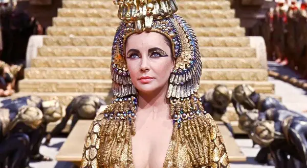 Elizabeth Taylor jako Kleopatra w złotej, bogato zdobionej sukni i koronie, z poddanymi klęczącymi u stóp.