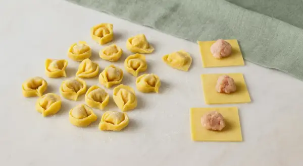 Frische Tortellini und Teigquadrate mit Füllung auf Marmorplatte.