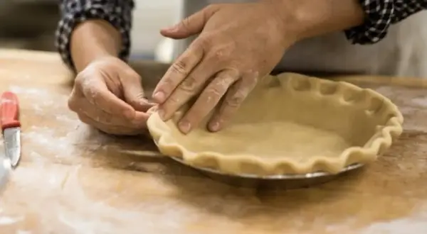 Mains farinées façonnant une pâte à tarte dans un moule. L'ajout d'une touche d'ammonium bicarbonate assure une croûte légère et croustillante.