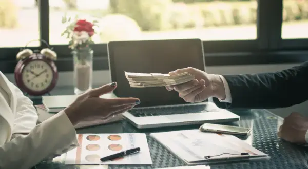 Dłoń przekazuje plik banknotów, co stanowi potwierdzenie przyjęcia gotówki. Na stole leżą dokumenty, laptop i telefon.