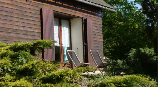 Chalet en bois avec étang de pêche privé en Franche-Comté. Deux chaises longues sur la terrasse, entourées de verdure luxuriante.