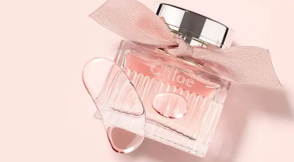 Szklana buteleczka L'eau de Chloe z różową wstążką i kroplami perfum na delikatnym, różowym tle.