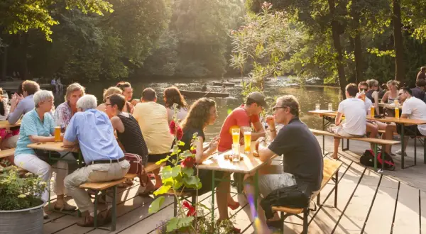 Menschen genießen das Leben im cafe am neuen see. Sie sitzen an Holztischen, trinken Bier und unterhalten sich. Im Hintergrund fließt ein ruhiger Fluss.