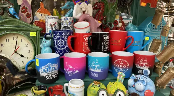 Kolekcja kubków z napisami "Sopot" i morskim motywem, idealne pamiątki z wakacji. W tle zegar i ceramiczne figurki.