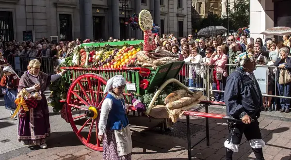 Carro con ofrenda de frutos, pan y verduras, adornado con flores, desfila entre una multitud.