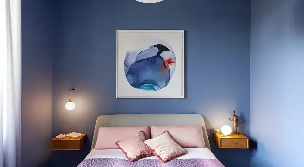 Camera da letto con pareti blu intenso e un quadro astratto. Letti con cuscini rosa e una coperta viola creano colori rilassanti.