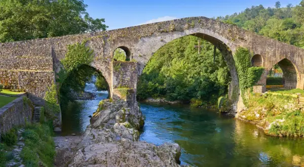El puente del Sella, de piedra y cubierto de vegetación, cruza un río de aguas verdes rodeado de árboles frondosos.