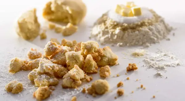 Knusprige Streusel mit gemahlenen Mandeln, bestäubt mit Puderzucker. Im Hintergrund Teig und Zutaten für eine köstliche Leckerei.