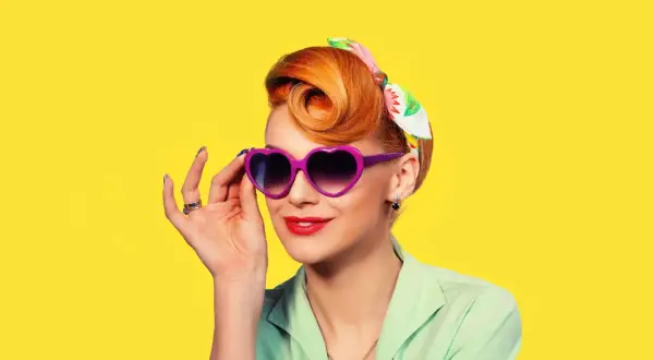 Une femme au style rockabilly, cheveux coiffés en chignon, porte des lunettes violettes en forme de cœur et un foulard coloré.