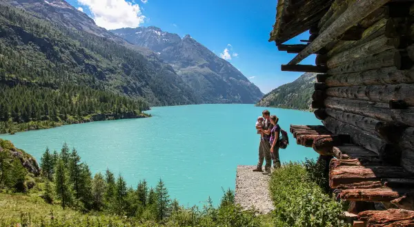 Un couple avec un bébé admirent le lac d'Aoste, entouré de montagnes verdoyantes et d'une cabane en bois rustique.