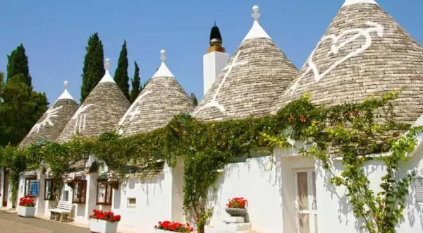 I trulli di Puglia, con i loro tetti conici e le decorazioni bianche, incantano sotto il cielo azzurro, avvolti dal verde rampicante.