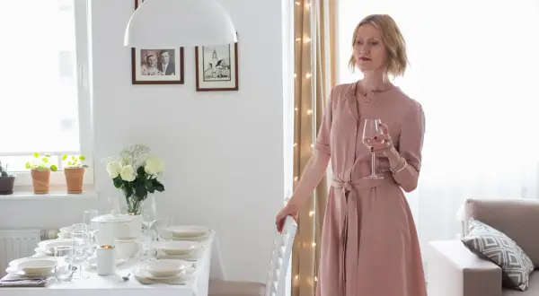 Elegancka kobieta w różowej sukience z kieliszkiem wina, przy nakrytym stole. Idealna stylizacja co na wesele.