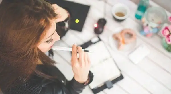 Kobieta pracuje przy laptopie, notuje w zeszycie. Na stole kawa, pączek i kwiaty. Może to praca zdalna, która daje dodatek za prace w weekendy.