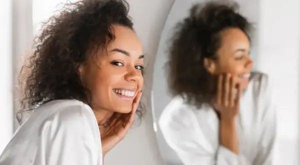Lächelnde Frau mit lockigem Haar, die sich im Spiegel betrachtet. Ihre Haut strahlt, vielleicht dank Retinol jeden Tag.