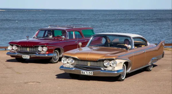 Två klassiska Plymouth Fury 1960, en röd kombi och en guldfärgad coupé, står vid havet.