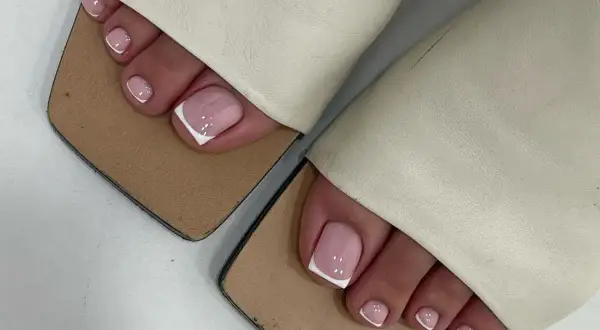 Elegancki pedicure french na różowych paznokciach stóp, ozdobionych białymi końcówkami i kropkami, w beżowych klapkach.