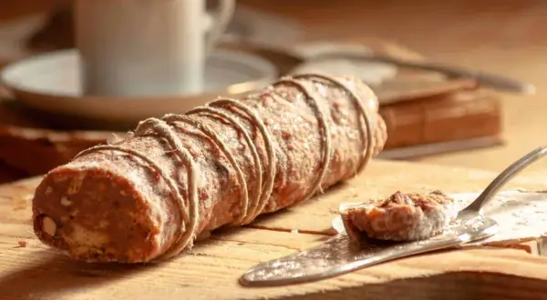 Ein rustikaler salame al cioccolato, gebunden mit Juteschnur, liegt auf einem Holzbrett. Daneben eine Kaffeetasse und ein Stück auf einem Tortenheber.