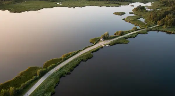Droga przecinająca jezioro, otoczona zielenią i lasem. W oddali mały domek, jakby cyców na wodzie.