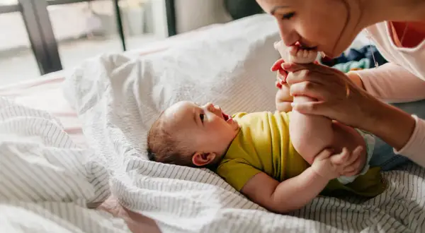 Mama całuje stópki niemowlaka, który leży na łóżku. Widać, że dziecko szybko rośnie, kiedy podwojenie masy urodzeniowej jest już za nim.