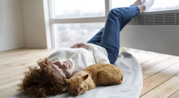 Mujer sonríe con su gato naranja sobre una manta, disfrutando del calor de la aerotermia en un piso.