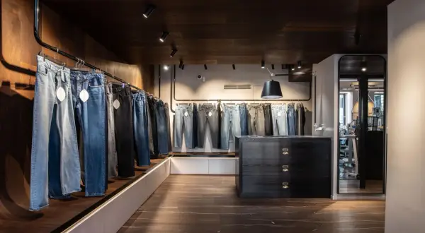 Jeans appesi in un negozio, con una **gamba affusolata** che si nota in primo piano. L'ambiente è moderno, con pavimenti in legno scuro e illuminazione a faretti.
