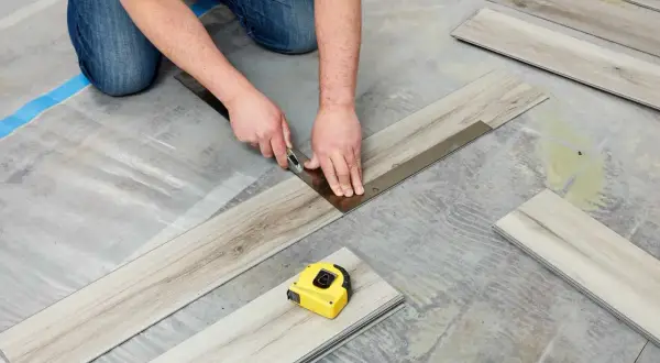 A person is cutting vinyl plank flooring that is not laying flat during installation. A tape measure is nearby.
