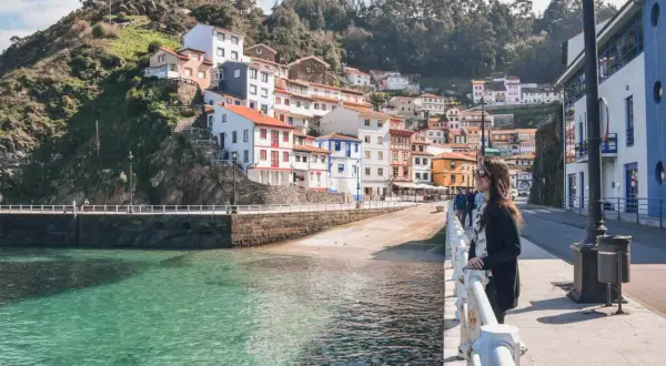 Una mujer admira la costa de **pueblos de Asturias cerca de Galicia**. Casas coloridas se aferran a la ladera verde junto a un mar turquesa.