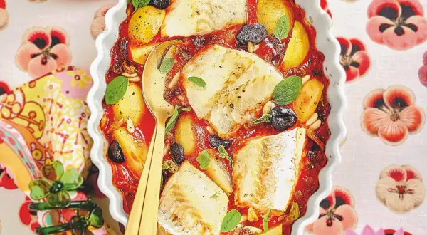 Piatto di baccalà alla romana in teglia con pomodoro, patate, olive e pinoli, guarnito con basilico fresco.