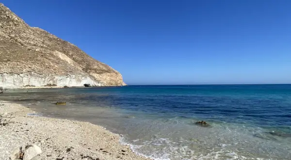 La **playa Peñón Blanco** se extiende bajo un cielo azul intenso. Las olas acarician la orilla de guijarros, mientras una imponente montaña rocosa se alza a la izquierda, con una cueva marina visible en su base.