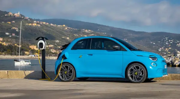 Abarth EV azul cargando junto al mar, con un velero y colinas al fondo.