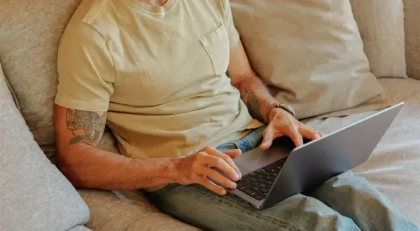 Man med tatuering på armen sitter i soffan med laptop, kanske för att han är hemma under en ny sjukperiod.