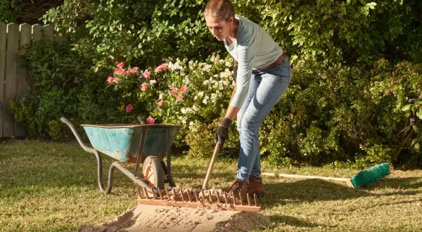 Frau verteilt mit Harke Sand auf dem Rasen. Eine Schubkarre und ein Besen liegen daneben.