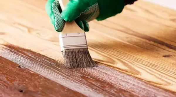 Mano con guante verde aplicando barniz a madera. Proceso para saber como impermeabilizar madera y protegerla.