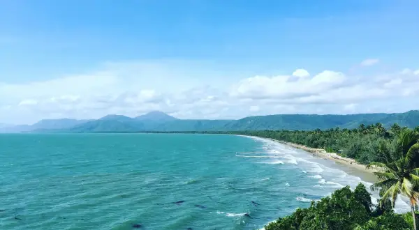Vacker kustlinje vid Port Douglas med turkost hav, grönskande berg och en gyllene strand under en molnig himmel.