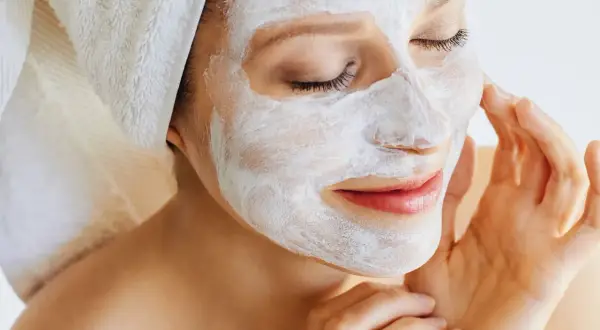 Frau mit weißer Maske im Gesicht, die ihre Haut pflegt. Ein Handtuch ist um ihren Kopf gewickelt.