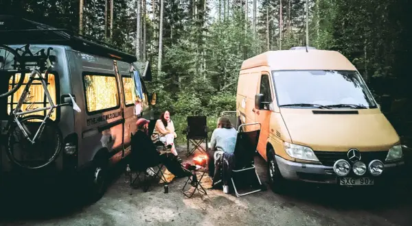Två husbilar parkerade i skogen, med folk som sitter runt en lägereld. En cykel sitter på takräcket på den ena bilen. En härlig bild av van life.