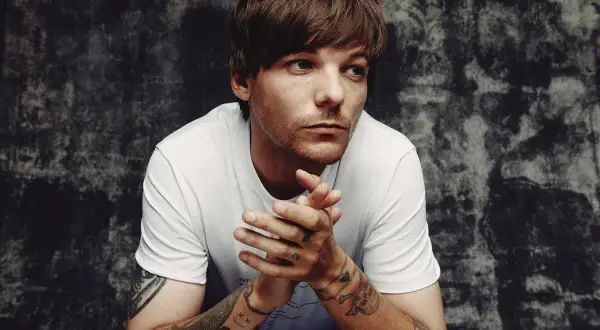 Louis Tomlinson zamyślony, z dłońmi złożonymi przed sobą. Fani czekają na jego nadchodzące wydarzenia.