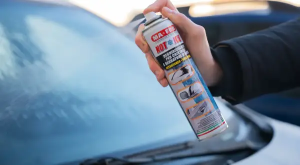 Mano che spruzza un prodotto per togliere il ghiaccio dal parabrezza dell'auto.