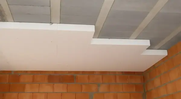 Ocieplenie stropu styropianem w trakcie budowy. Białe płyty styropianowe montowane pod betonowym stropem, nad ceglanymi ścianami.