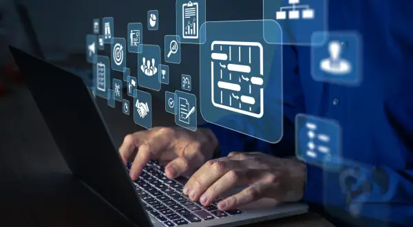 Dłonie pracują na laptopie, nad którym unoszą się ikony symbolizujące integrację systemów, zarządzanie projektami i współpracę.