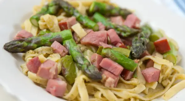 Pyszne tagliatelle ze szparagami i kawałkami szynki, podane na białym talerzu.