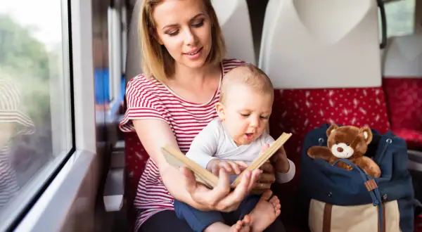 Mama czyta książeczkę rocznemu dziecku w pociągu. Obok siedzi plecak z pluszowym misiem.