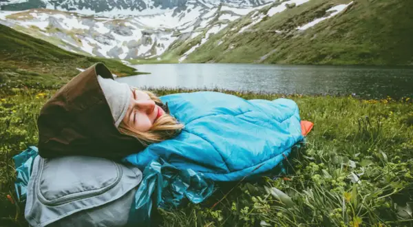 Persona durmiendo en saco azul, con mochila gris, en un prado verde junto a un lago y montañas nevadas. Vivac acampada perfecta.