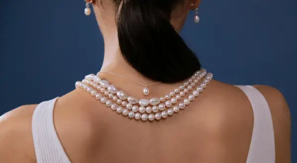 Kobieta z tyłu w białej bluzce, ozdobiona kilkoma naszyjnikami z pereł i kolczykami. Miara pereł podkreśla jej elegancję.